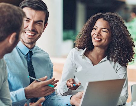 A group of people in professional attire having a meeting, smiling, and discussing with papers and laptops on the table.