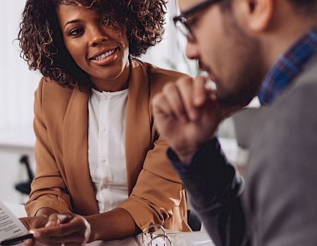 Two people are discussing something, with one holding a paper. They appear to be in an office setting, engaged in conversation.