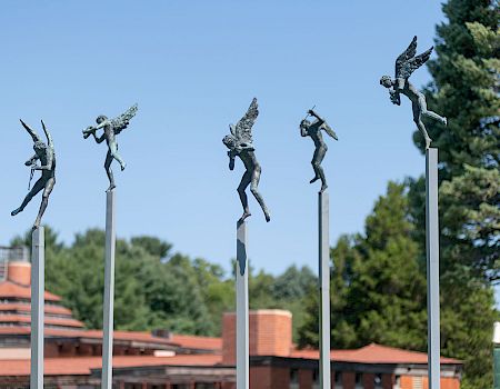 Sculptures of angels playing instruments are perched on tall poles against a backdrop of trees and a building with a red roof.